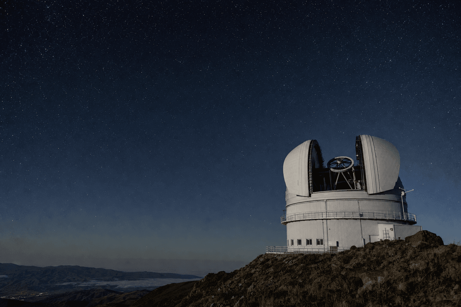 A mountaintop astronomical observatory at night, its dome open to a sky full of stars above a dark ridgeline.